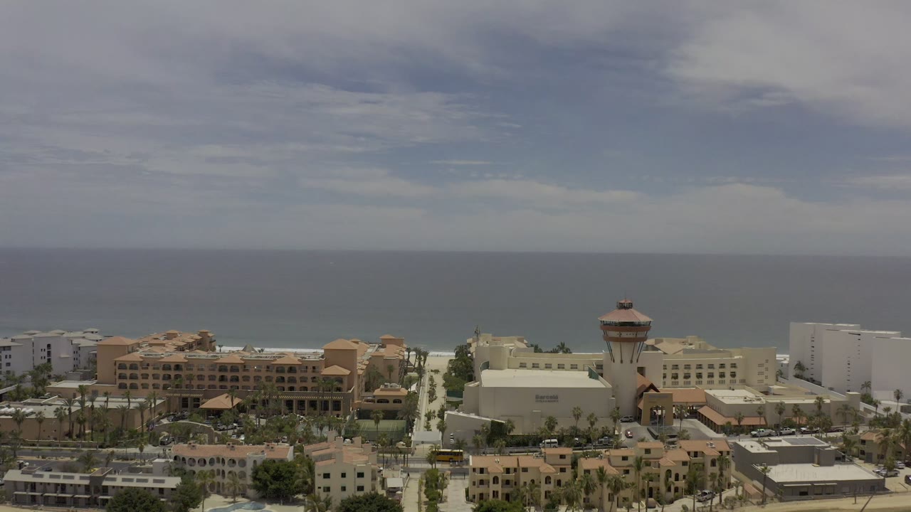 A Los Cabos aerial view holds on beachfront development, with the tower slightly right of center, pale surf line, broad ocean, clouded sky, and no visible people.