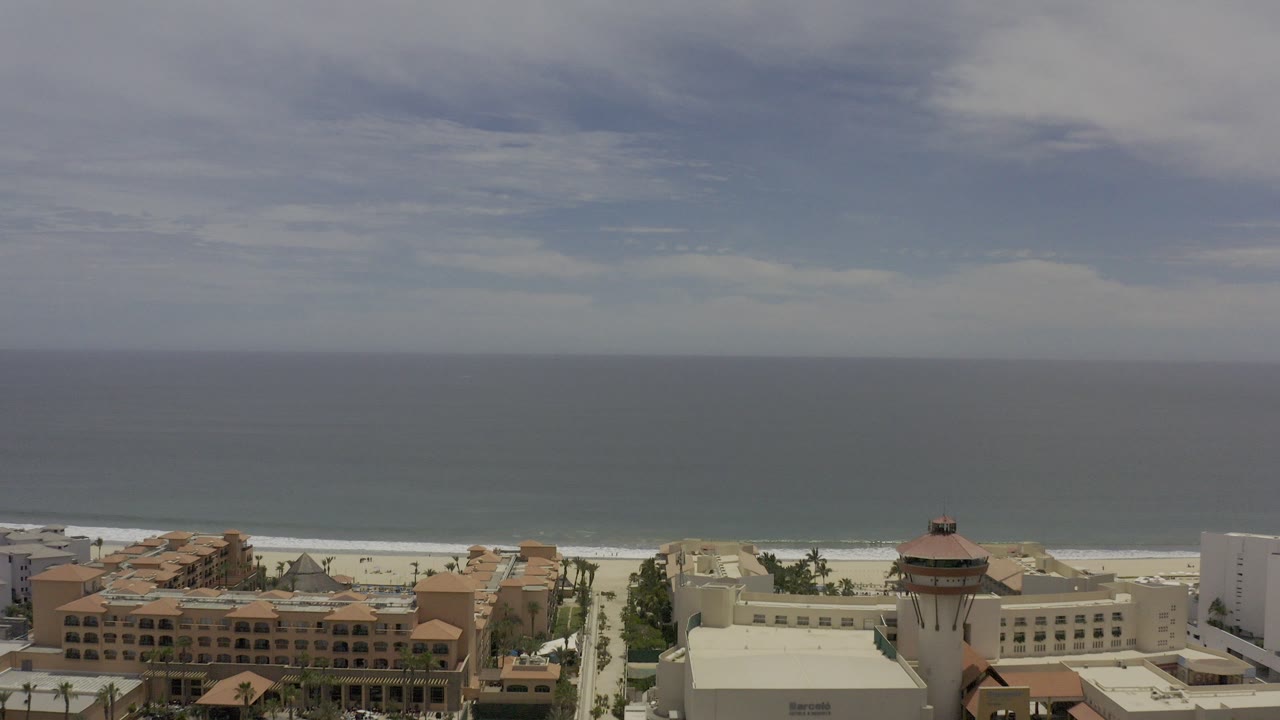 Opening aerial frame over Los Cabos, Mexico, with hotel roofs at the bottom, a pale beach and surf line beyond, blue-gray ocean to the horizon, and no visible people.