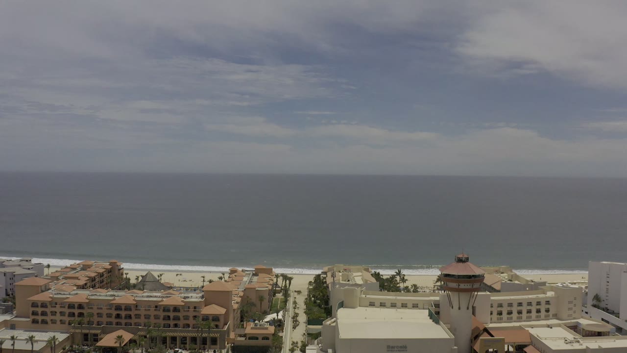 Aerial Los Cabos coastline view with more resort buildings spanning the lower edge, a thin white surf band, wide ocean, hazy horizon, and no visible people.