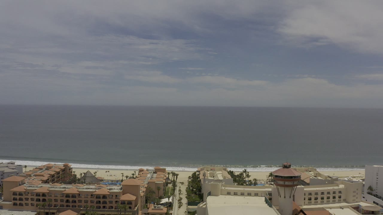 Beachfront resort roofs below a wide ocean and cloudy sky.