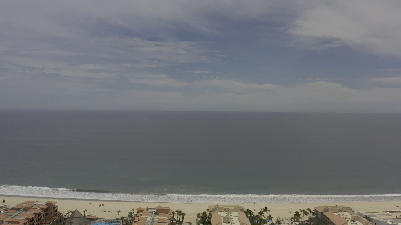 Wide Los Cabos beach and ocean view with beachfront rooftops at the bottom edge, surf along the sand, a distant horizon, partly cloudy sky, and no visible people.