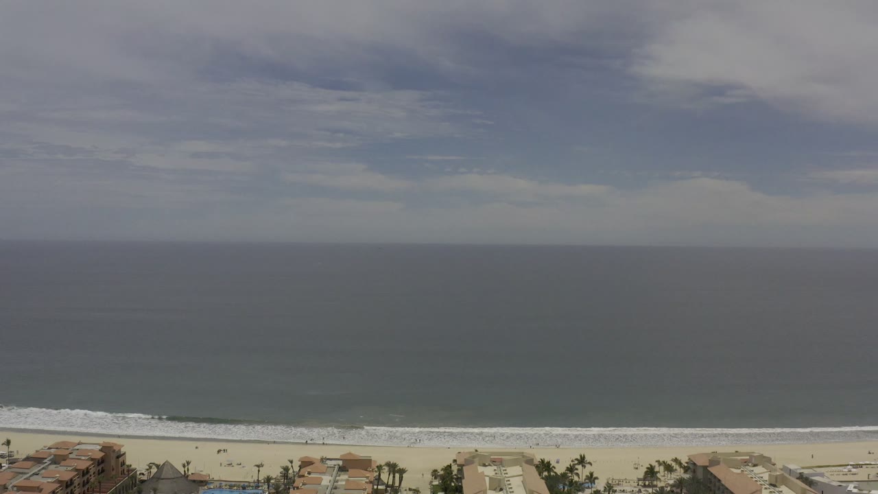 Elevated Los Cabos coastal frame with pale sand, a white surf band, broad blue-gray water, beachfront structures near the lower edge, and no visible people.