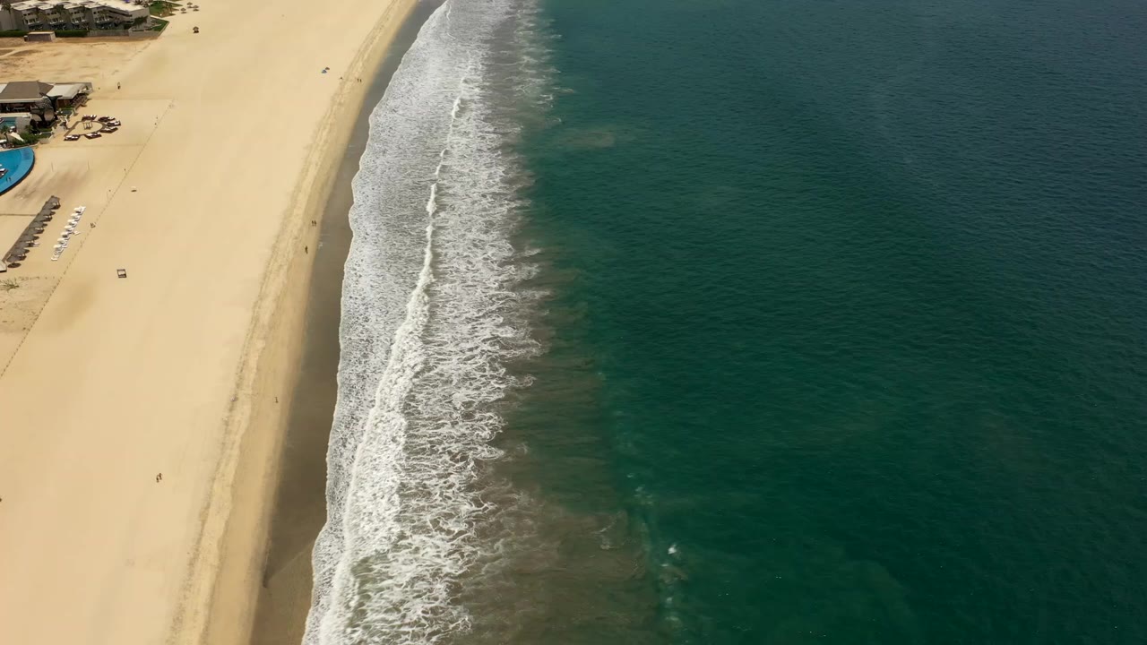Aerial view over a Los Cabos beach with pale sand at left, white surf along the shoreline, green ocean at right, beachfront houses near the top edge, and tiny people visible on the sand.