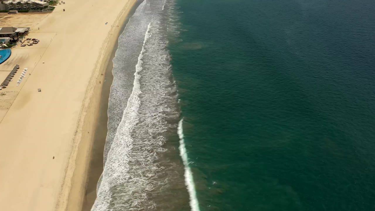 Aerial frame of Los Cabos coastline with a broad strip of sand, foamy waves curving down the shore, green water to the right, beach furniture near the left edge, and small people visible on the beach.