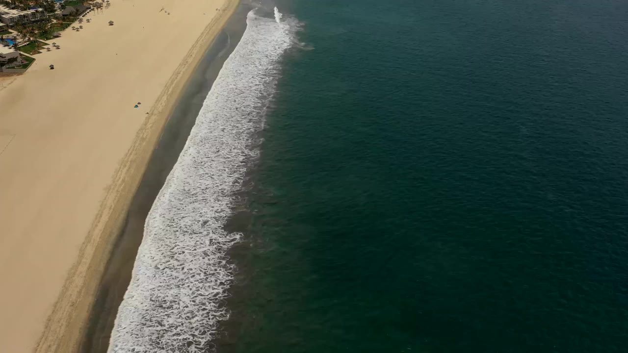 Aerial frame from Los Cabos looking down on a diagonal shoreline, with pale sand and faint tracks on the left, breaking white waves in the middle, open dark water on the right, and no visible people.