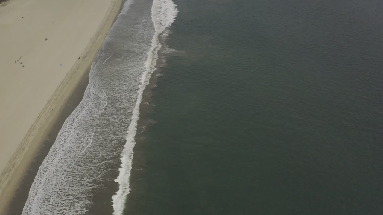 A Los Cabos shoreline fills the frame with tan sand on the left and a band of foamy waves pushing along the darker water, with no visible people.