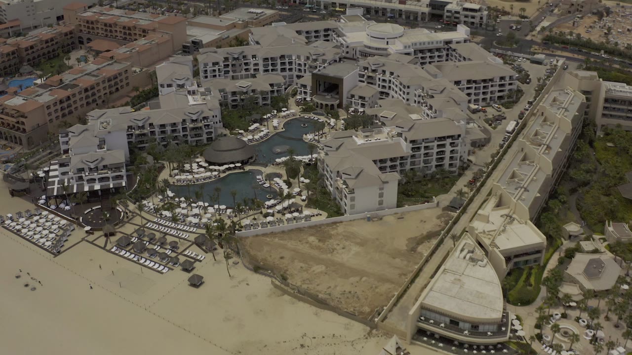 A slightly shifted overhead view of the Los Cabos resort shows the central pool area, rows of lounge chairs near the sand, neighboring buildings, and no visible people.