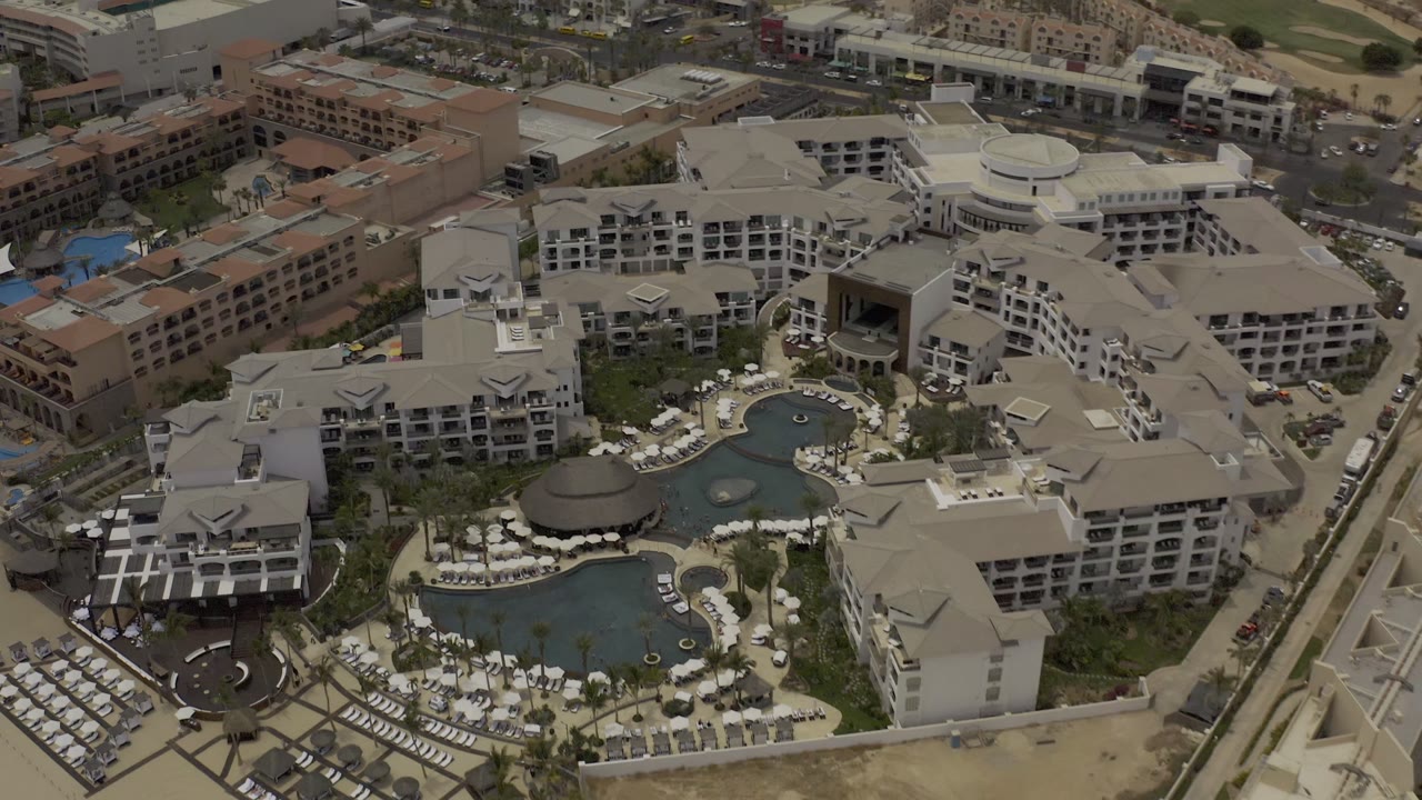 Aerial frame in Los Cabos showing a central resort pool area surrounded by hotel wings, white umbrellas, palm trees, access roads, and no visible people.
