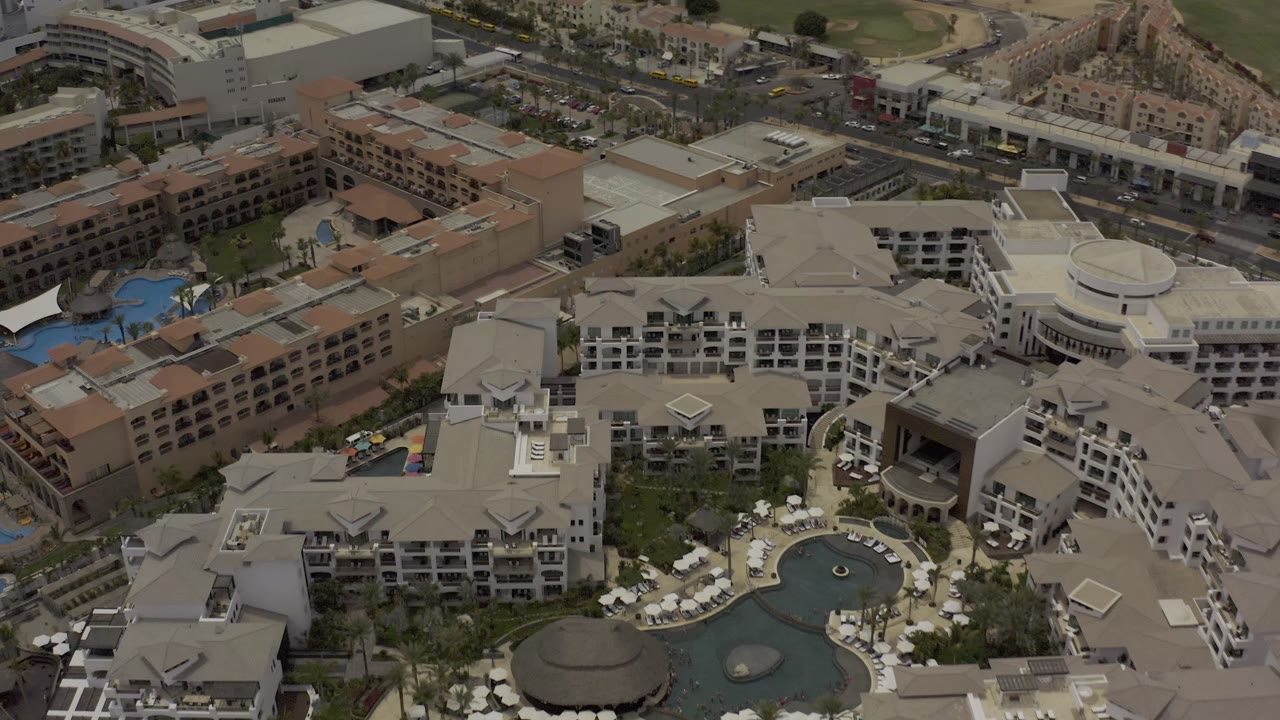 Aerial view in Los Cabos showing resort rooftops, blue pool sections, landscaped courtyards, road traffic, and no clearly visible people.