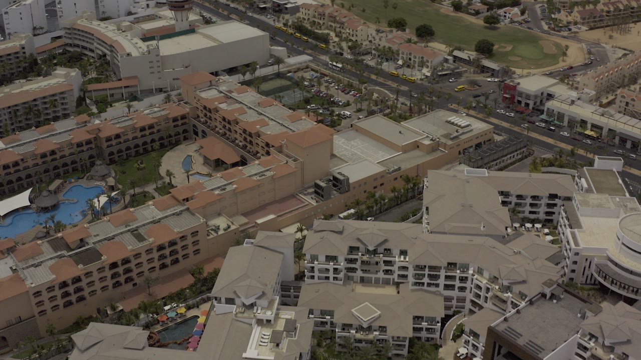 Overhead view in Los Cabos, Mexico showing tan and white rooftops, a curved road, parked vehicles, and a blue pool area, with no visible people.