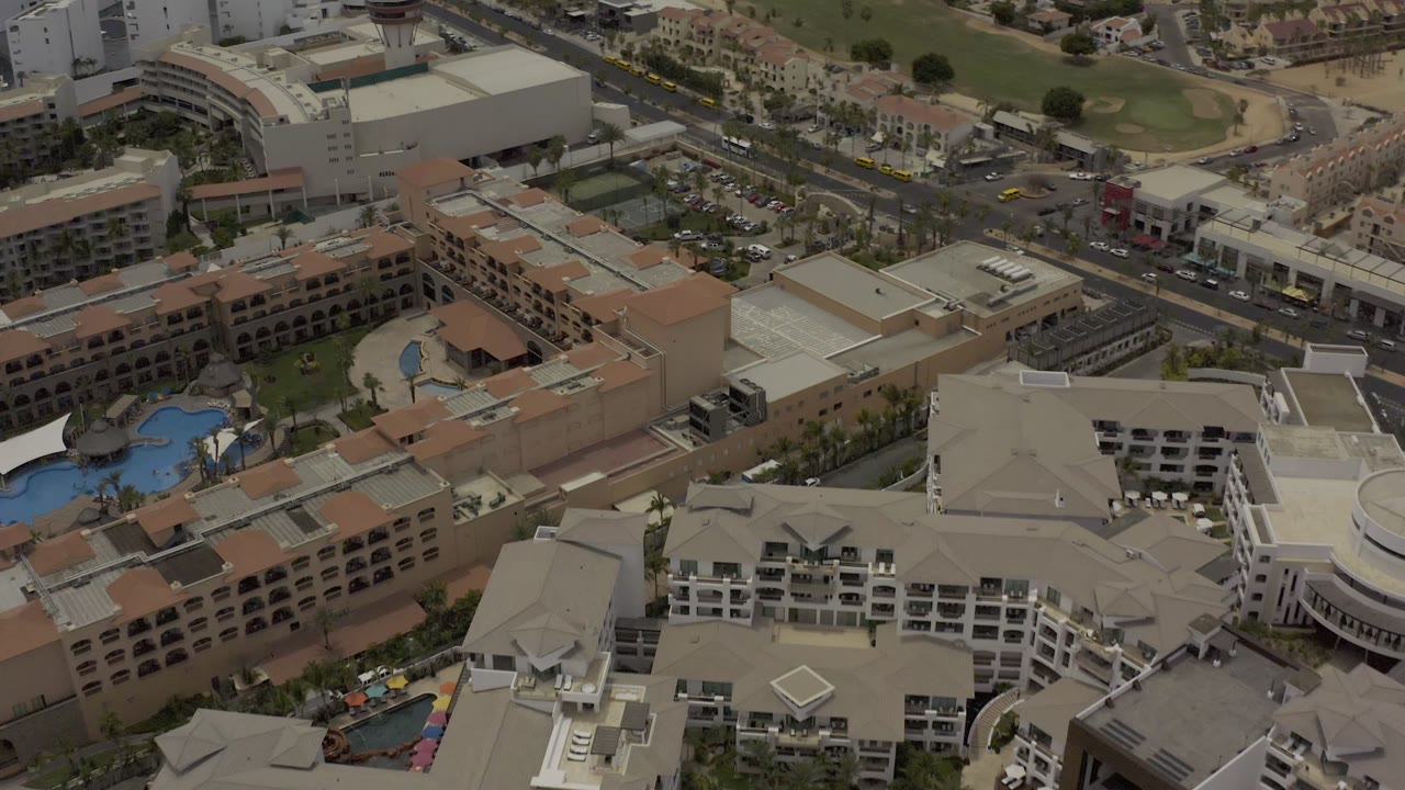 High aerial frame over Los Cabos buildings with a blue pool at left, parking areas, intersecting roads, and no visible people.