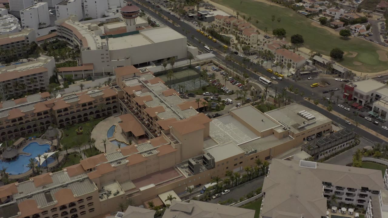 Aerial frame in Los Cabos showing resort rooftops, tennis courts, curved pool areas, busy roads, tall white buildings, and no visible people.