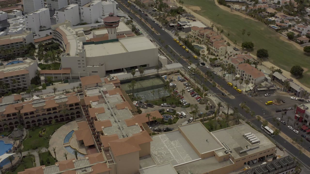 Los Cabos aerial view with tan-roofed resort structures in the foreground, light traffic on the central road, white hotel blocks beyond, and no visible people.