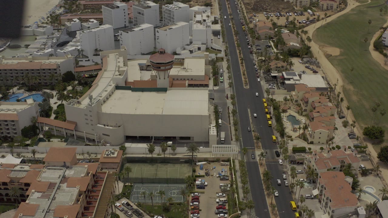 High aerial frame over Los Cabos, Mexico, showing white buildings, a divided road with cars, pools, red-tile roofs, and a green course area, with no visible people.