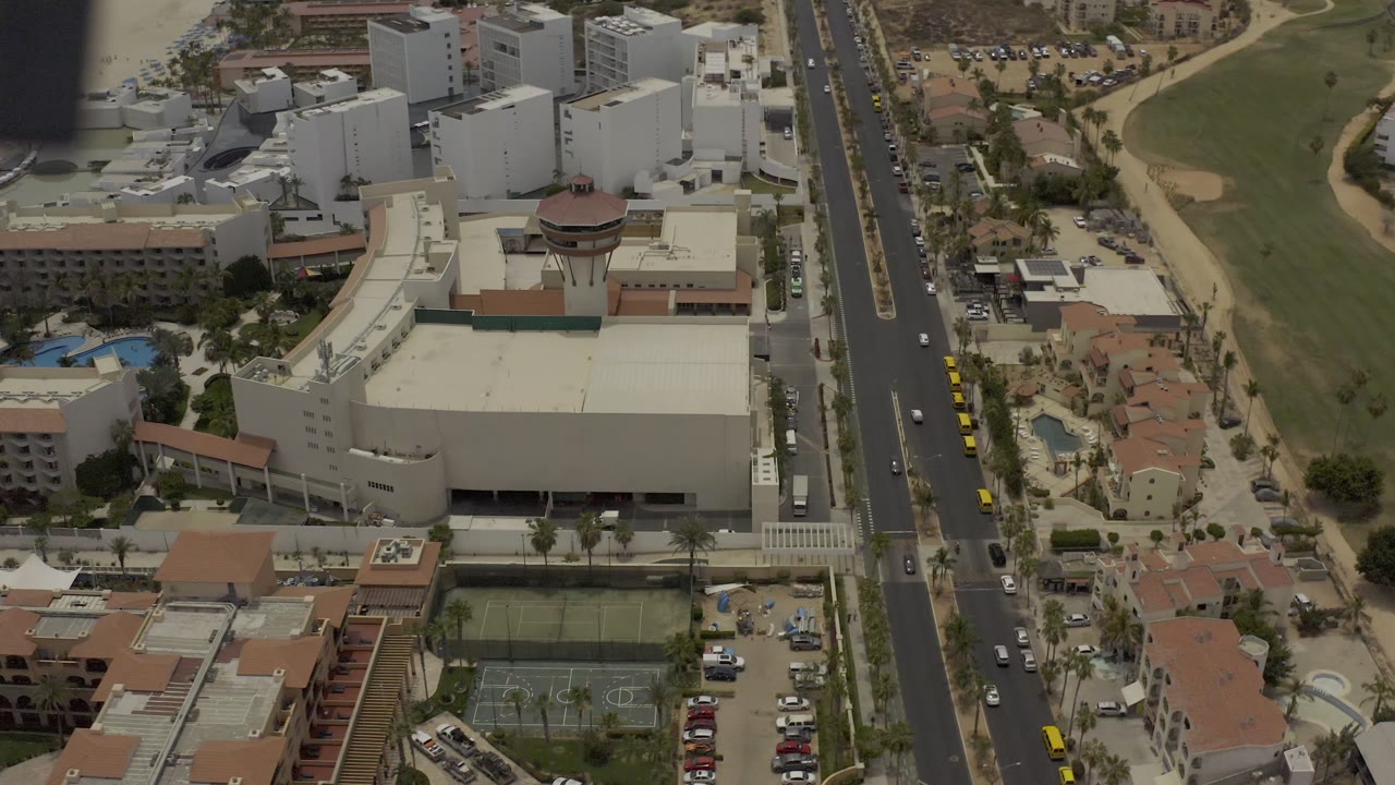 Aerial view in Los Cabos, Mexico, with a central road, pale commercial-style buildings, parked cars, tennis courts, small pools, and landscaped grass, with no visible people.