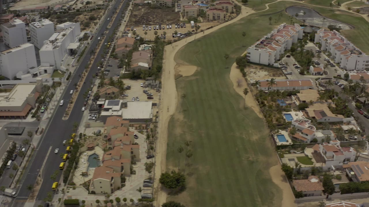 Los Cabos aerial frame with the green field centered, highway traffic along the left side, apartment blocks at the top, and no visible people.