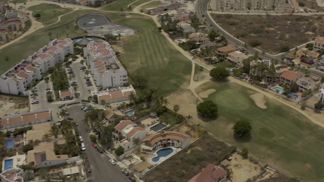Los Cabos aerial view with the residential cluster shifted toward center, showing fairway grass, sandy cart paths, palm trees, a curved pool, and no visible people.