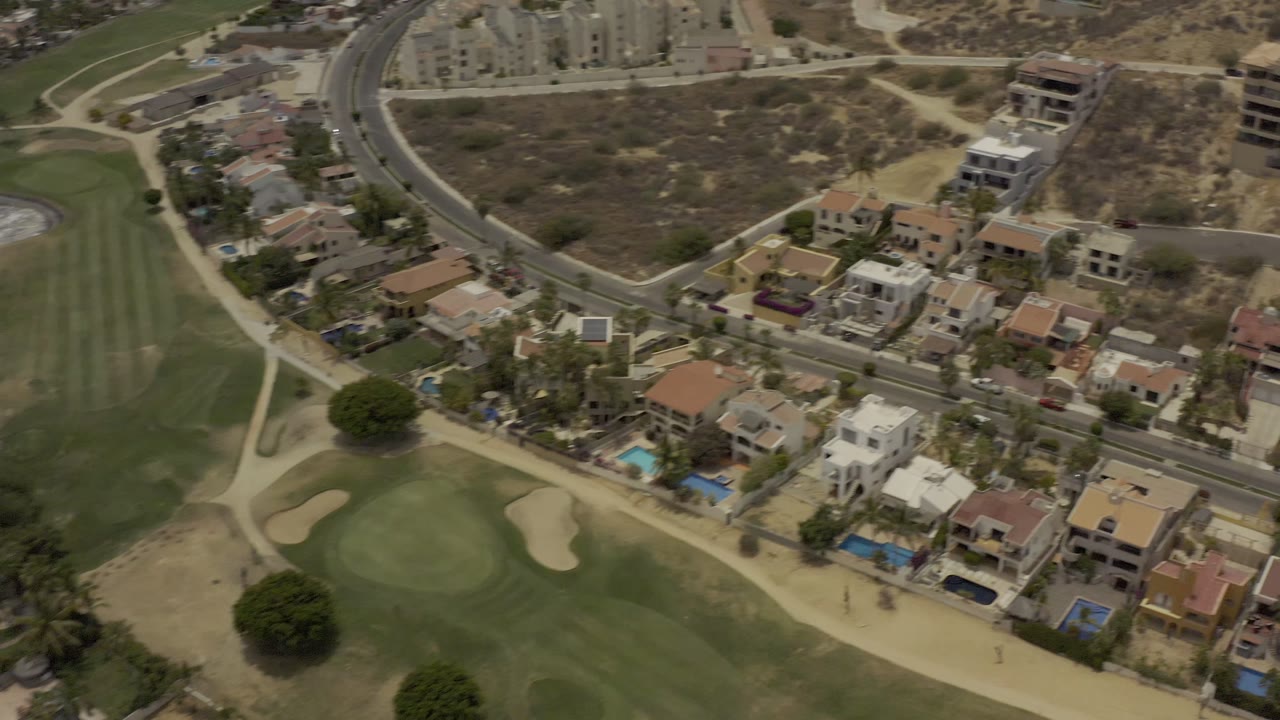 Aerial frame in Los Cabos, Mexico with a green fairway and cart path in the foreground, a row of houses with blue pools in the middle, and dry lots uphill, with no visible people.
