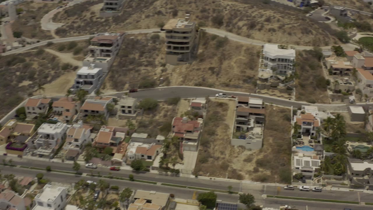 Aerial frame over Los Cabos, Mexico with villas, dry open lots, a paved hillside road, parked cars, and a bright blue swimming pool, with no visible people.