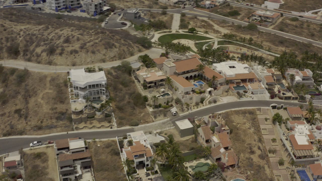 Aerial frame over Los Cabos showing hillside residential homes, curving roads, small pools, dry scrub terrain, and no visible people.