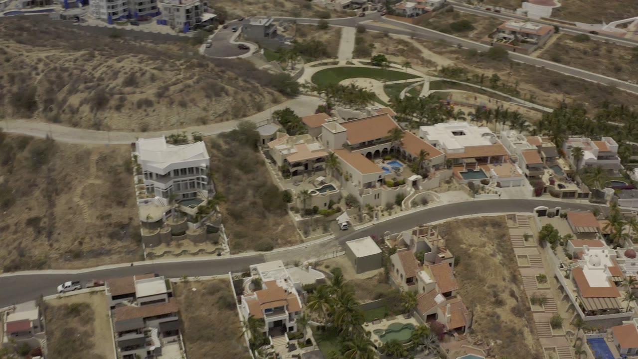 Aerial view in Los Cabos with villas along a curved street, dry hillsides above them, a small landscaped green area, and no visible people.