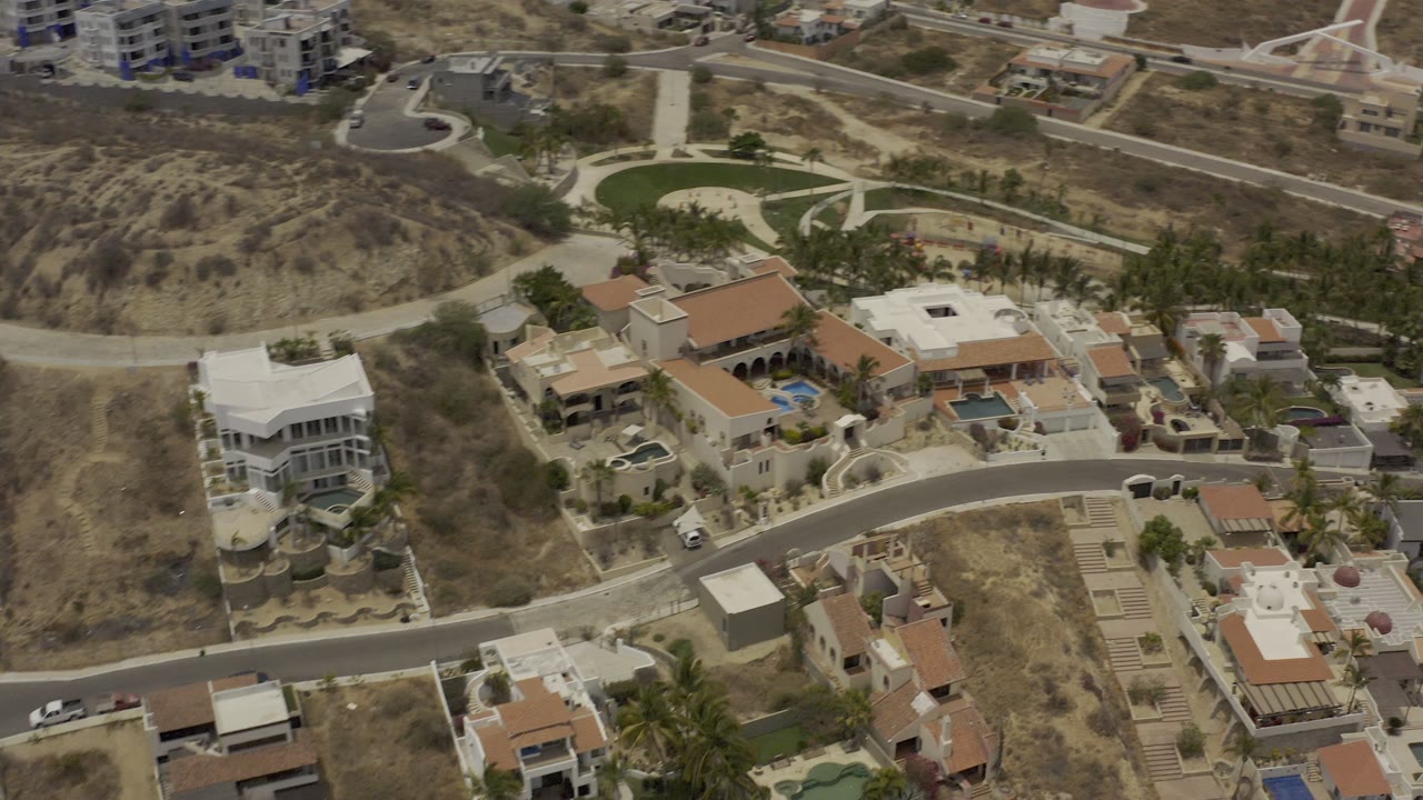 Top-down Los Cabos residential frame showing tile-roof houses, pale walls, blue pools, dry undeveloped lots, and no visible people.