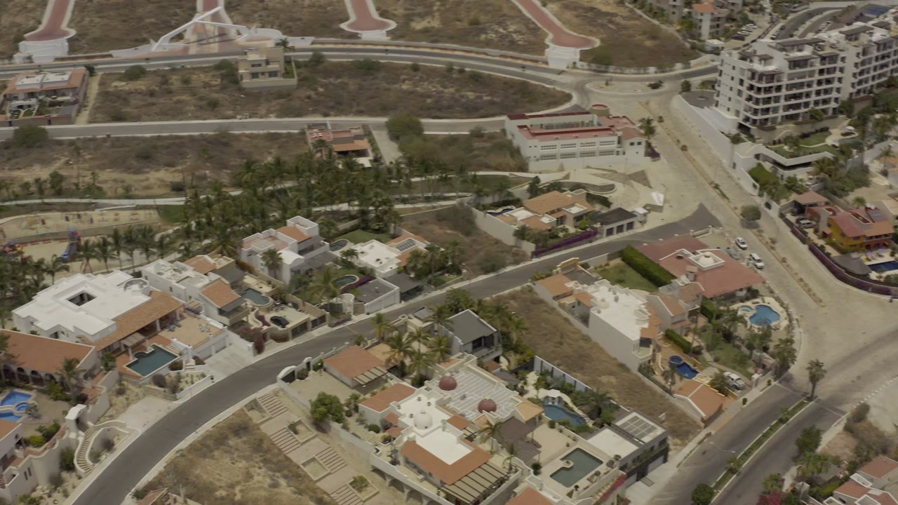 Aerial view in Los Cabos showing a curved residential road, clustered villas, blue pools, palms, dry open land, construction areas, and no visible people.