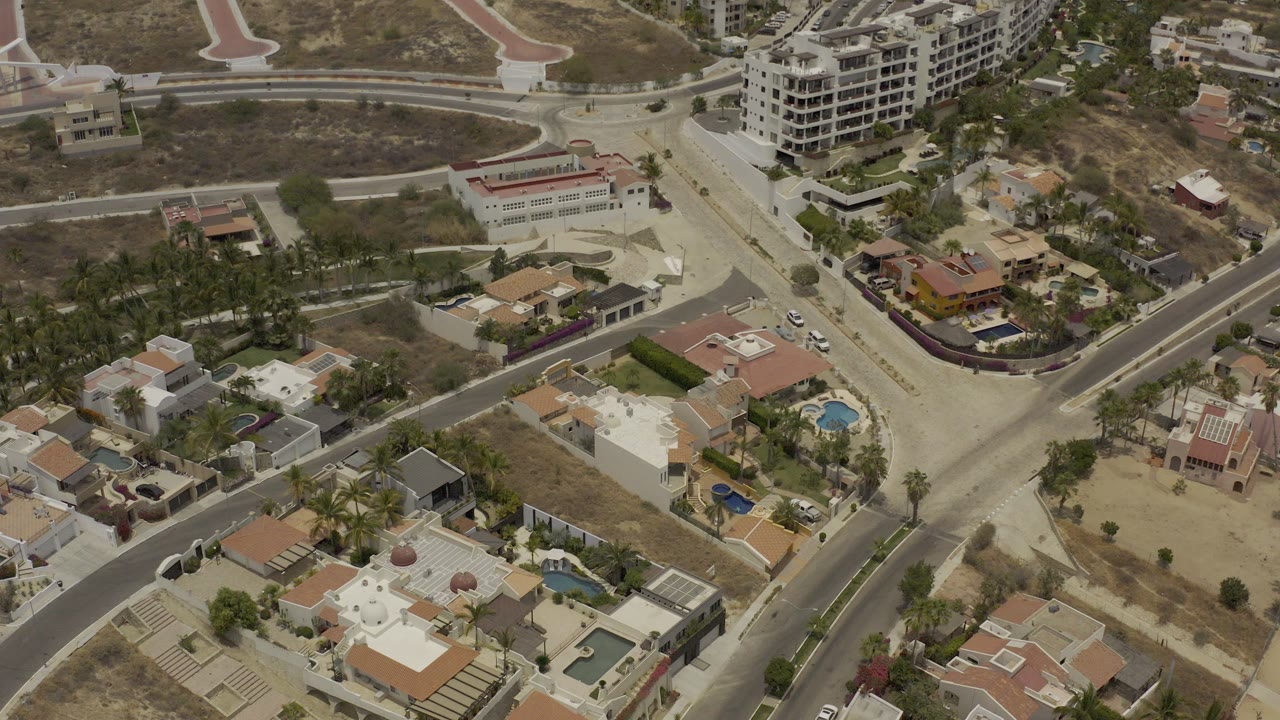 High aerial view over Los Cabos, Mexico, showing curved roads, private homes, blue pools, dry lots, and mid-rise buildings, with no visible people.