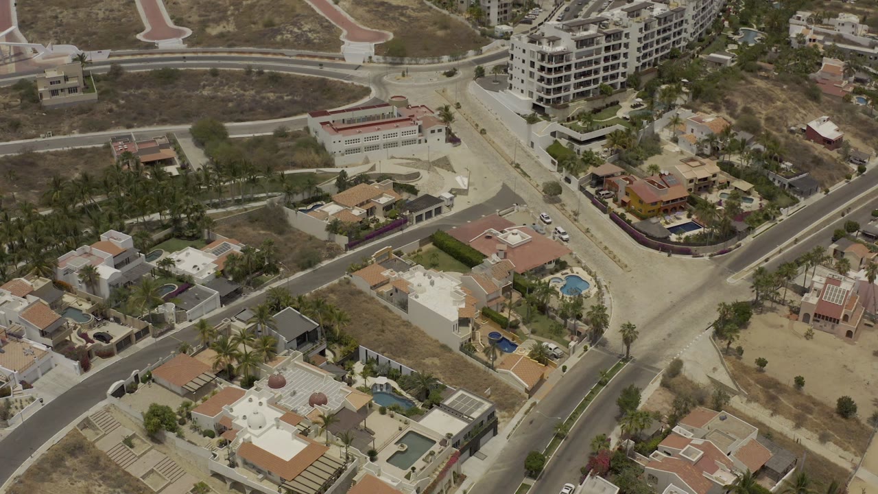 Los Cabos aerial frame with a light-colored road junction, nearby houses, palm landscaping, pools, and unfinished lots, with no visible people.