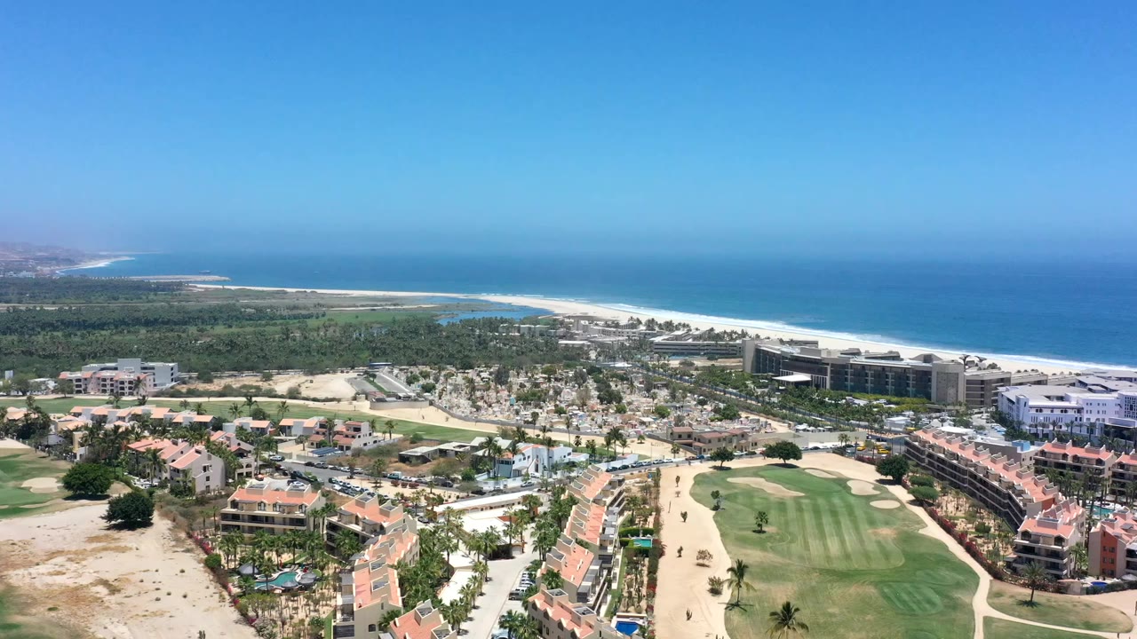 High aerial view over Los Cabos with red-roof buildings, green golf fairways, a white beach, blue ocean, and no visible people.