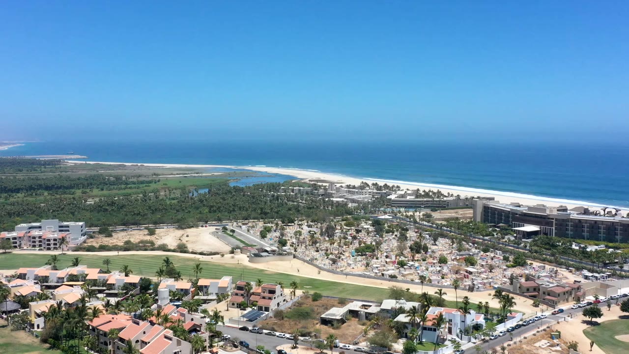 Wide aerial view in Los Cabos, Mexico, showing the town grid below, a curved coastal inlet, white beach, deep blue sea, and no visible people.