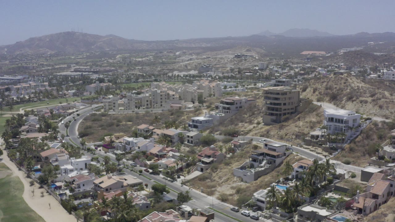 Opening aerial frame over residential Los Cabos, Mexico, with hillside houses, a curving road, dry terrain, distant mountains, and no visible people.