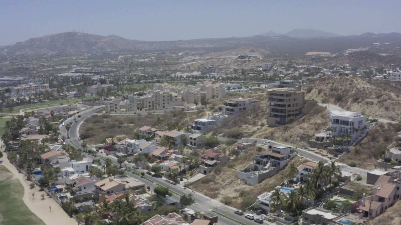 Aerial frame over Los Cabos homes and streets, with a rounded dry hill near the center, larger buildings beyond it, and no visible people.