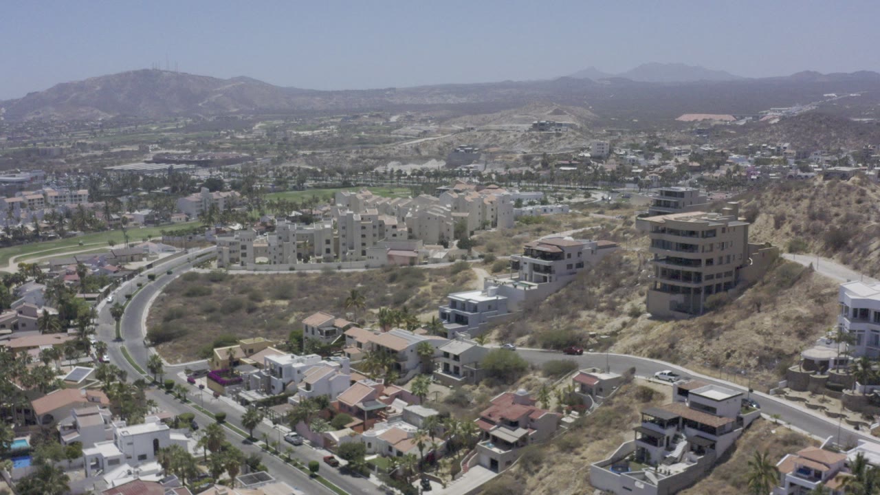 Aerial frame over Los Cabos, Mexico showing curving roads, hillside homes, apartment blocks, dry terrain, distant mountains, and no visible people.