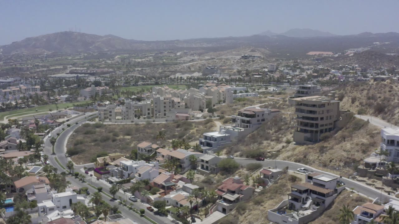 Aerial view in Los Cabos, Mexico with foreground rooftops, a winding road, beige apartment buildings, dry slopes, distant mountains, and no visible people.
