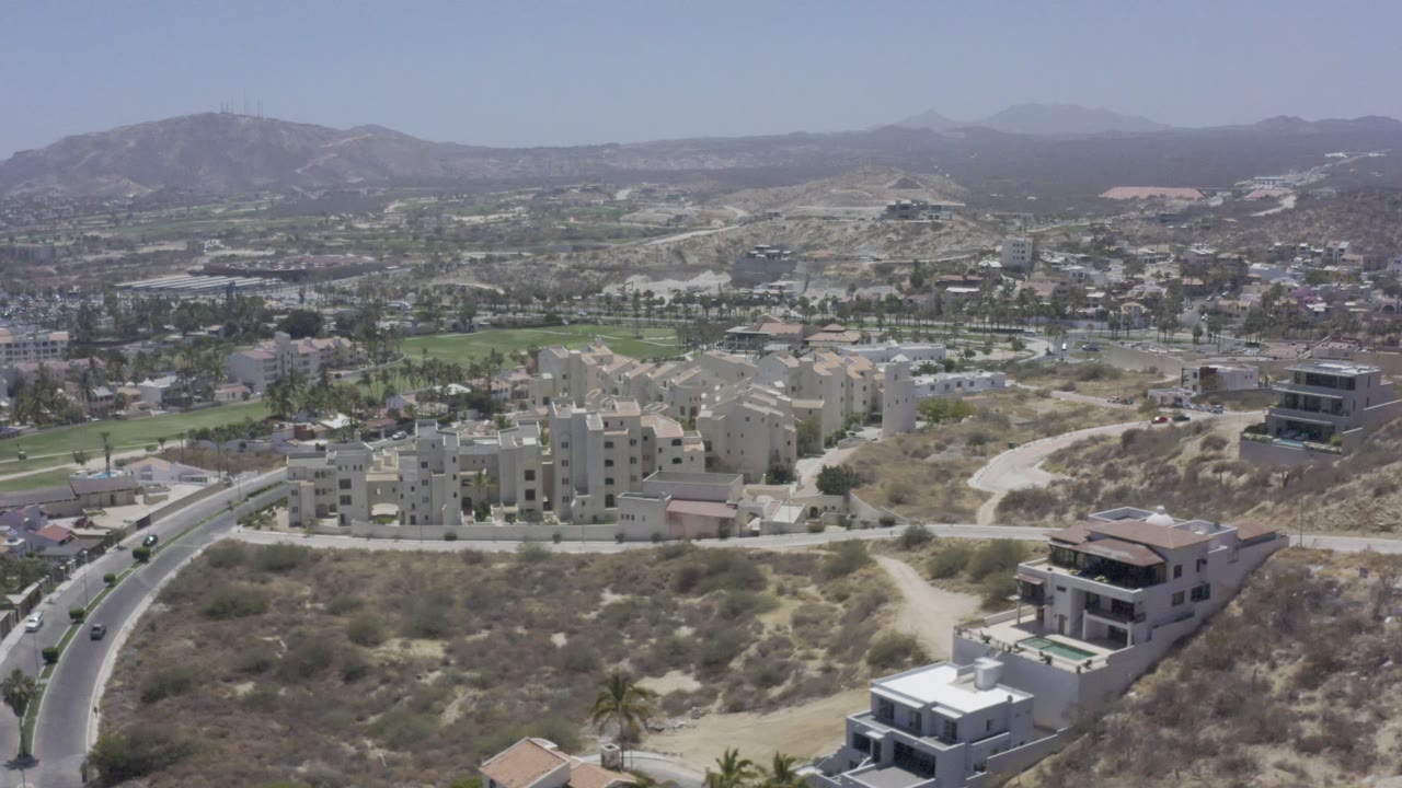 Aerial view in Los Cabos, Mexico, looking over apartment buildings near a curved road, scattered houses on dry terrain, green strips, distant hills, and no visible people.