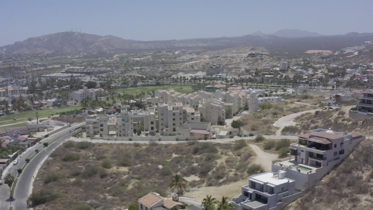 Aerial Los Cabos frame with clustered apartment blocks centered below dry hills, a curved road at left, modern homes at right, and no visible people.