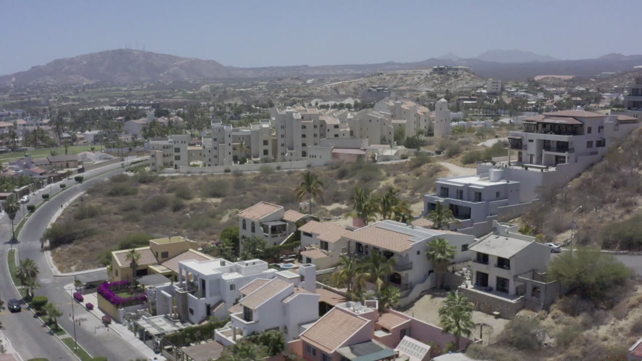 Los Cabos aerial frame with a road bending through the left side, clustered white and beige homes, scrubby hillside terrain, palms, mountains, and no visible people.