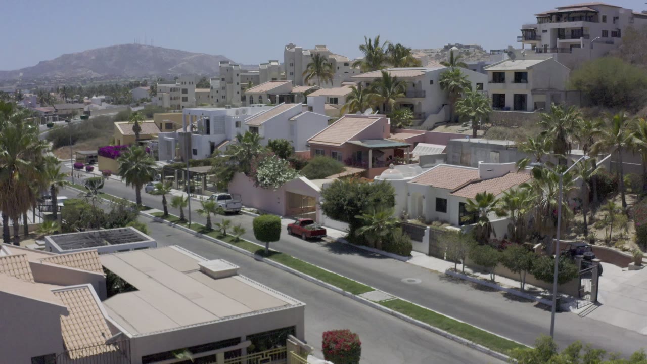 At 0:01, Los Cabos houses and palms sit above a quiet residential road with parked cars, dry hills in the background, and no visible people.