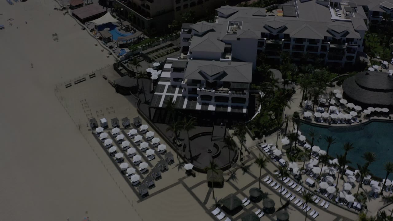 Aerial view in Los Cabos with rows of empty beach loungers on sand, resort buildings across the center, and a blue pool area at the right with no visible people.