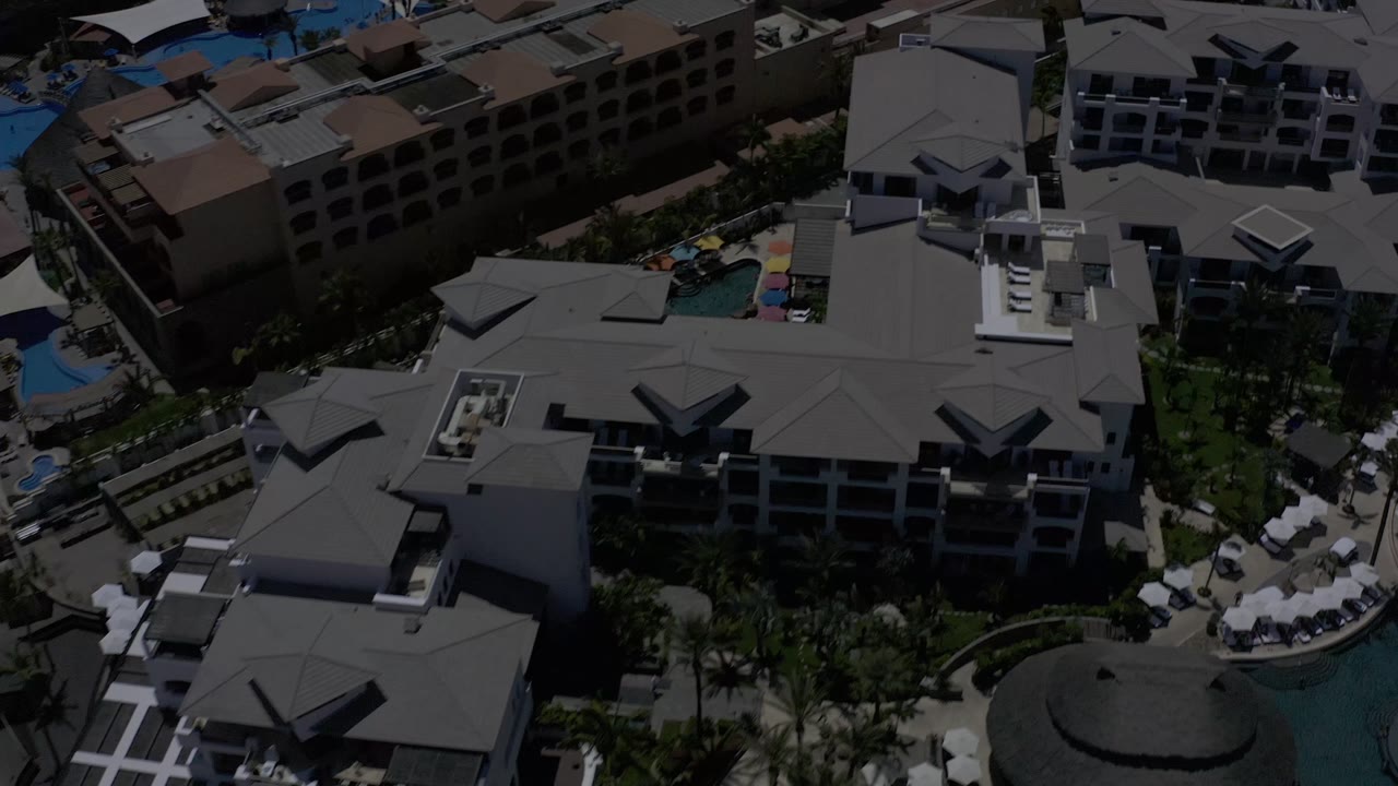 Los Cabos resort rooftops fill the frame, with a curved pool edge, shaded umbrellas, balconies, and no visible people.