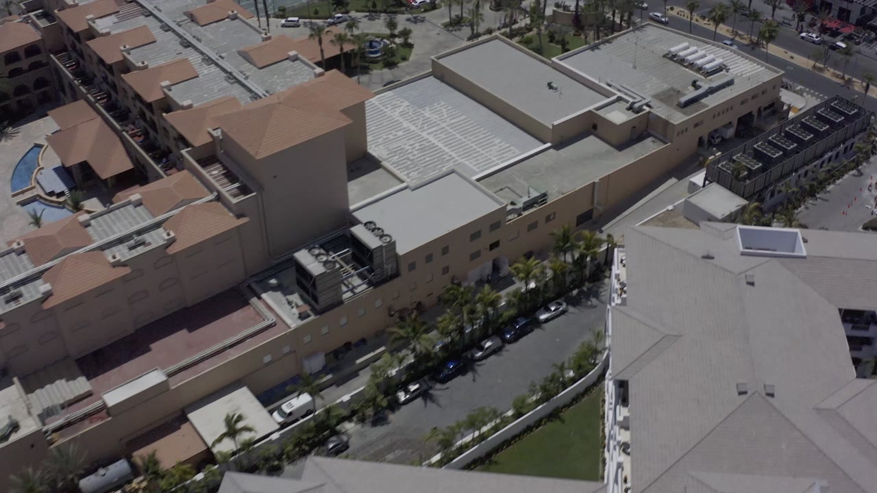 A second aerial moment in Los Cabos, Mexico, shows the same mall block from slightly farther across the roofline, with parked cars along the curb and no visible people.