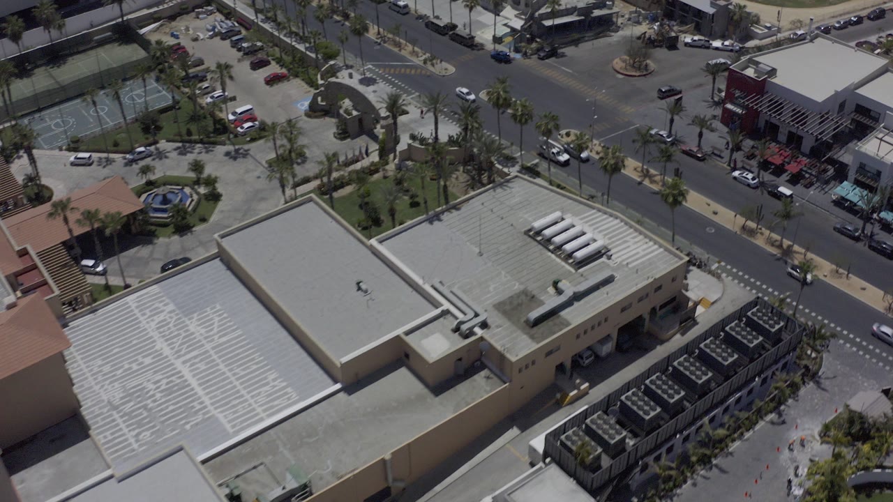 Aerial frame over Los Cabos, Mexico showing flat-roofed commercial buildings, a palm-lined road, parked cars, and no clearly visible people at the start of the clip.