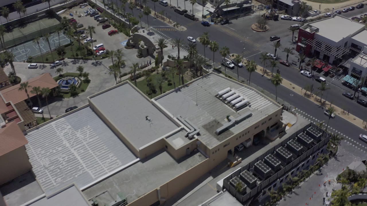 Aerial frame over Los Cabos, Mexico with a large pale rooftop in the foreground, road lanes beyond it, palm trees, a small landscaped plaza, and no clearly visible people.