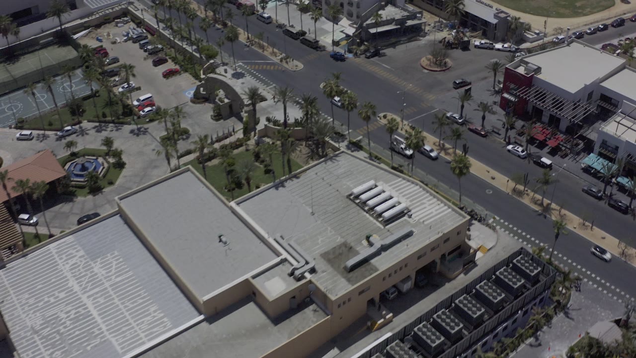 Overhead street view with a large flat roof, parked cars, palms, and crosswalk markings.
