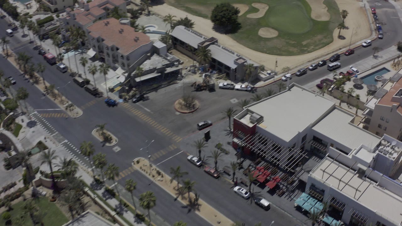 Aerial frame over Los Cabos showing a road intersection, low buildings, parked cars, palm trees, and golf greens in the background, with no visible people.