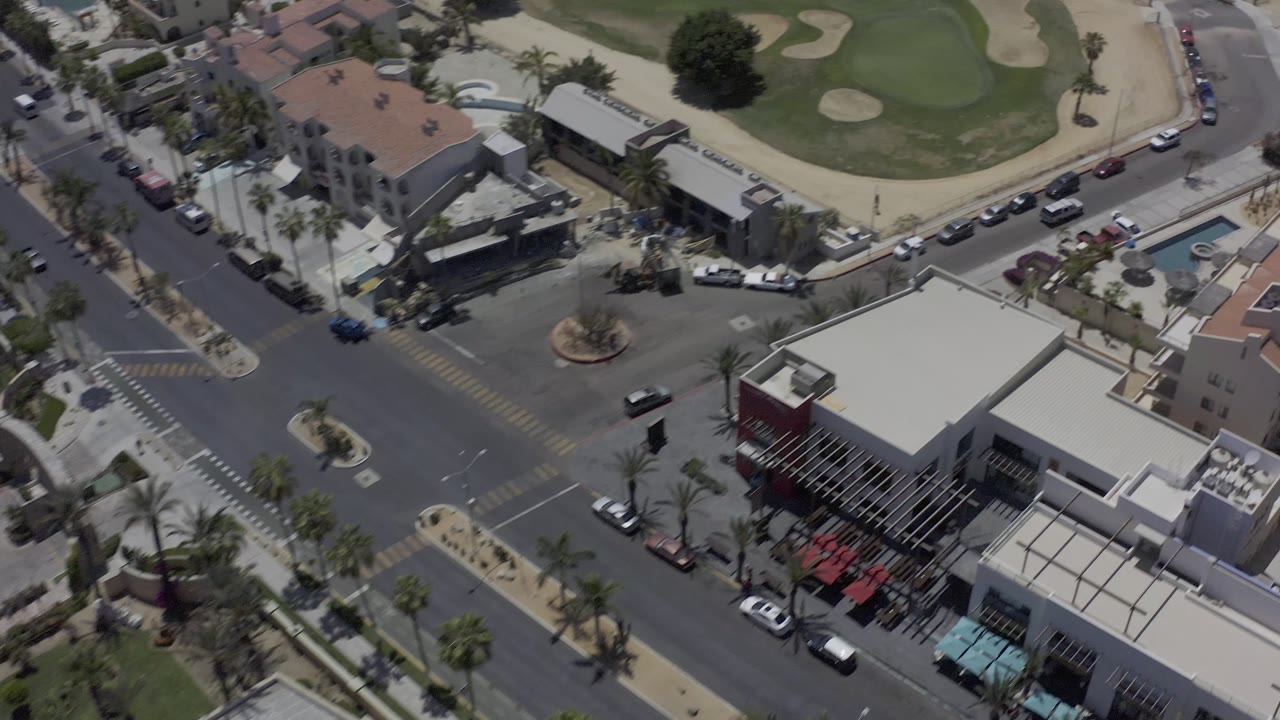 High drone view in Los Cabos with the intersection lower left, commercial rooftops near center, parked cars along curbs, and golf fairways behind them, with no visible people.