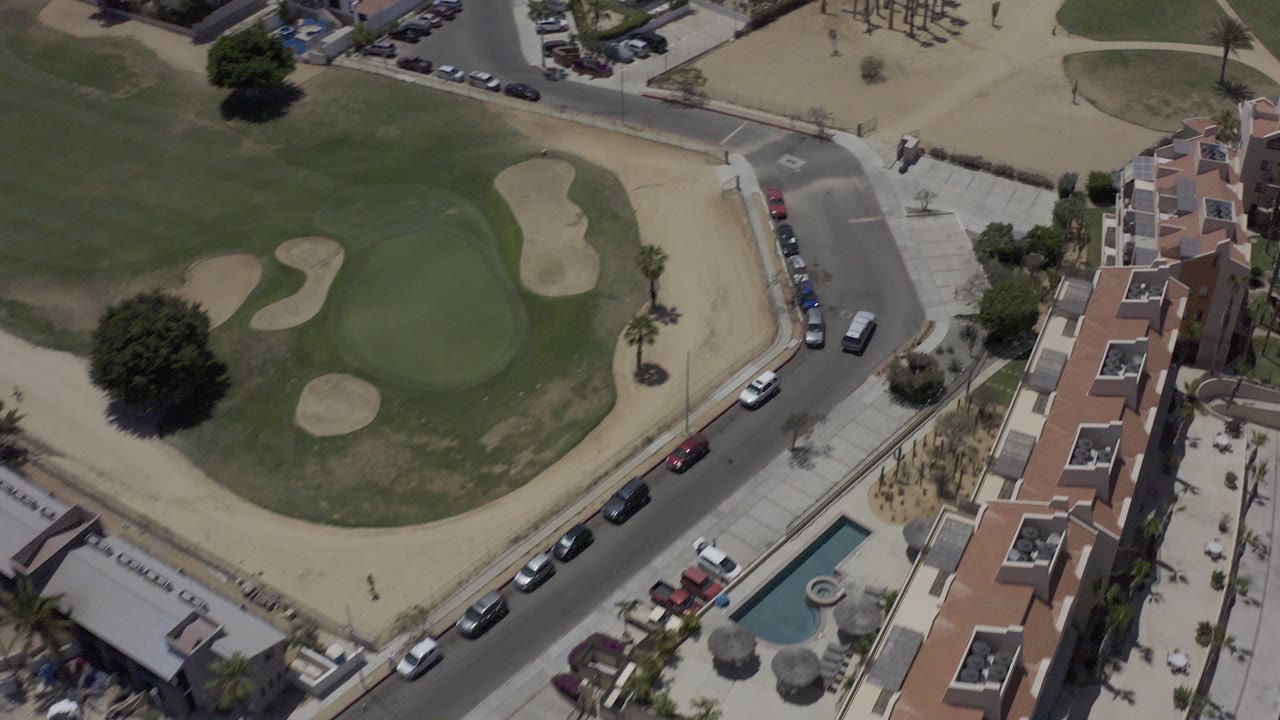 Overhead frame in Los Cabos, Mexico showing a green field with sand traps, tan-roofed housing, parked cars along a curved street, and no visible people.