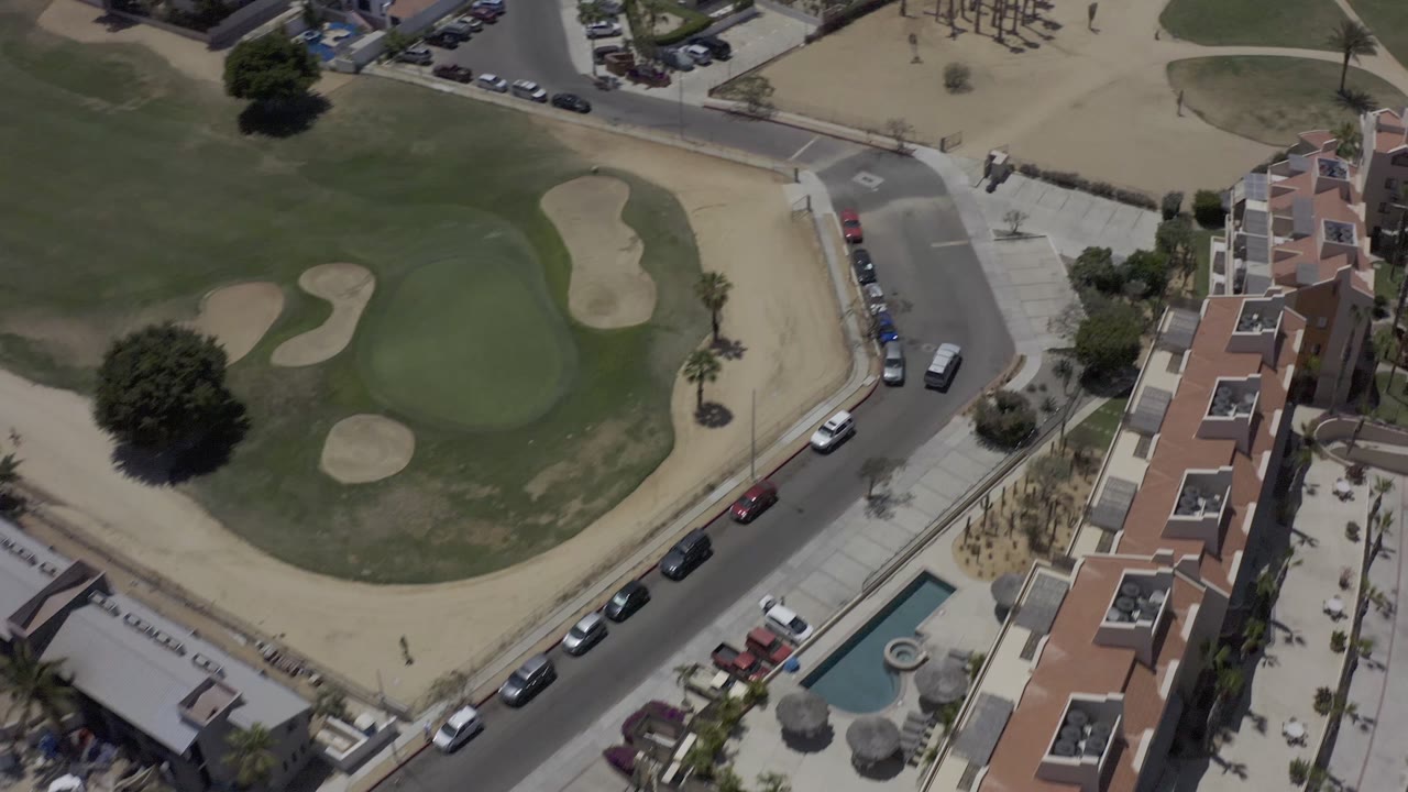 Aerial view in Los Cabos, Mexico with the residential building row more centered beside green turf, sand traps, street parking, and no visible people.