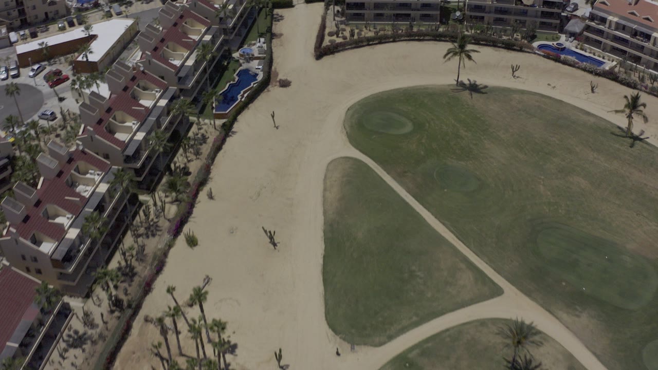 Overhead view of green field, pale paths, buildings, pools, and palm trees.